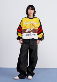 Sweatshirt oversized amarelo e preto riscado com gráfico de ciclismo, combinado com calças largas pretas. Ténis brancos com acentos roxos.