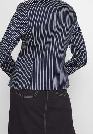 Marineblauw blazer met witte verticale strepen, gedragen met een zwarte rok met witte stiksels en achterzakken.