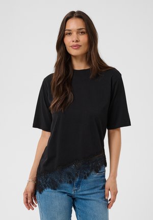 Femme aux longs cheveux bruns portant un haut noir à manches courtes avec bordure en dentelle et un jean bleu, debout devant un fond blanc uni.
