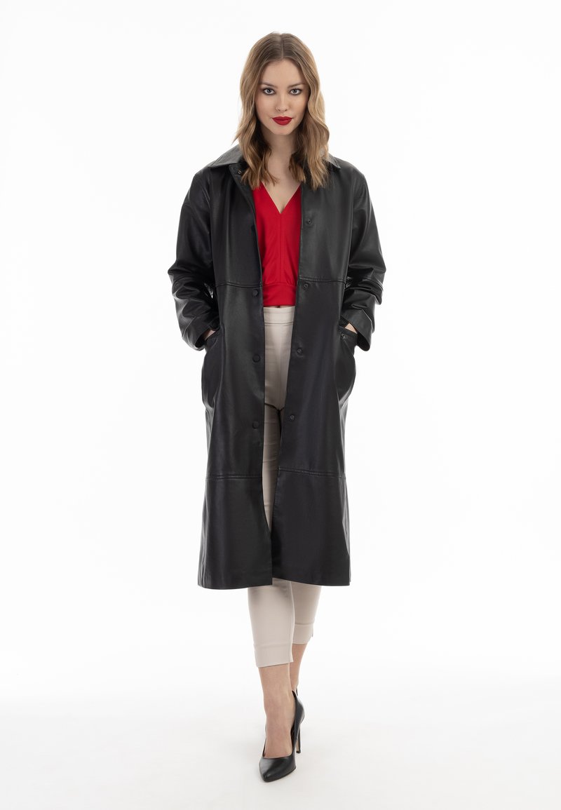 Cappotto lungo in pelle nera con chiusura a bottoni, tasche laterali e colletto ampio, abbinato a una maglia rossa e pantaloni beige cropped.