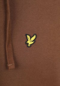 Bruine gebreide stof met een geel vogel-logo patch en een bijpassende bruine trekkoord aan de linkerkant.