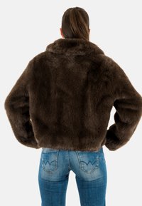 Giacca corta in finta pelliccia marrone con ampio colletto e maniche lunghe, vista da dietro, abbinata a jeans blu in denim.