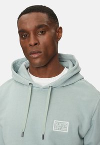 Sudadera azul claro hecha de tela suave, con un bolsillo frontal, cordones con detalles metálicos y un parche del logo bordado en el pecho.