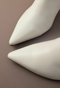 Chaussures blanches en cuir à bout pointu avec une texture lisse, présentant un design épuré et un style minimaliste, reposant sur une surface brune.