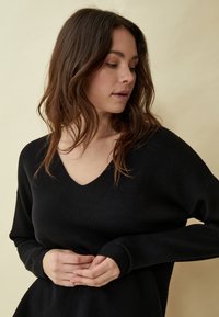 Črn pulover z V-izrezom, sproščenega kroja, dolgimi rokavi in rebrastimi manšetami. Mehek material in gladka tkanina, nošen do sredine telesa na nevtralni podlagi.