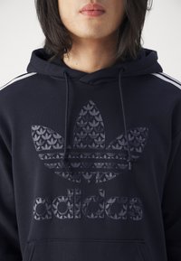 Navy blue hoodie featuring a textured logo and design with an embossed leaf pattern. Includes a front pocket and drawstring hood.