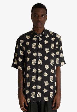 Giovane uomo che indossa una camicia nera a maniche corte con motivi astratti quadrati beige, in piedi davanti a uno sfondo bianco uniforme.