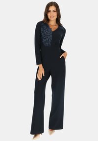 Granatowy kombinezon z długimi rękawami, z koronkowymi detalami w kwiatowy wzór na górnej części przodu. Dopasowany krój z szerokimi nogawkami i bocznymi kieszeniami.