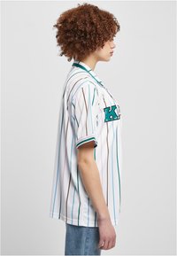 Bijela prugasta nogometna majica s tirkiznim, smeškim i ružičastim linijama, s kragnom i kratkim rukavima. Prikazuje slovo "K" u tirkiznoj boji.