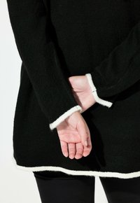 Černý pletený svetr s bílým lemováním na manžetách a lemu. Dlouhé rukávy, volný střih a texturovaný materiál. Ruce jsou umístěny za zády.