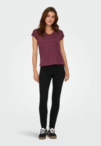 Femme aux cheveux bruns ondulés portant un haut maroon à manches courtes, un pantalon skinny noir et des baskets noires avec des lacets blancs, se tenant devant un fond blanc.