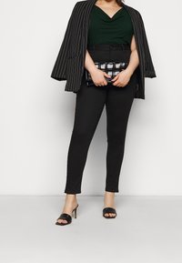 Svart pinnsträck blazer över en grön cowl-neck topp, svarta åtsittande byxor, svarta klacksandaler, håller en svart texturerad clutch, minimal bakgrund.