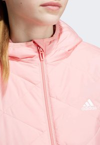 Chaqueta acolchada rosa claro con capucha y cremallera de longitud completa. Presenta un logo blanco en el pecho y está hecha de un tejido sintético suave.