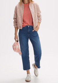 Różowa bomberka o fakturze, nałożona na rdzawe sweter, zestawiona z niebieskimi dżinsami o wysokim stanie. W ręku trzyma jasnoróżową czapkę w stylu tie-dye, do tego casualowe białe sneakersy.