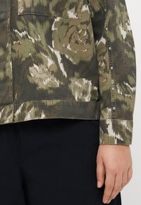 Chemise à motif camouflage dans des tons terreux de vert et beige, fabriquée en tissu texturé. Elle présente des boutons le long des poignets et une poche poitrine.