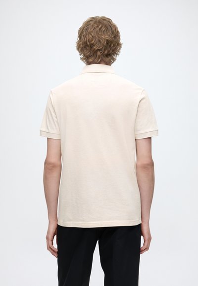 Personne aux cheveux bouclés châtain clair portant un polo beige à manches courtes et un pantalon noir, vue de dos sur fond blanc uni.