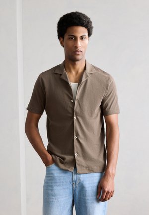 Mand med krøllet hår iført brun kortærmet skjorte med knapper over hvid top og lyseblå jeans, stående mod en ensfarvet baggrund.