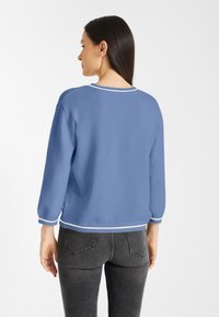 Blauwe gebreide trui met driekwartmouwen, met een ronde halslijn en witte bies bij de kraag en onderkant. G gedragen met donkere jeans.