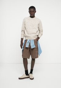 Man standing with hands in pockets, wearing beige sweater, brown cargo shorts, light blue shirt tied around waist, black socks, and beige sneakers.