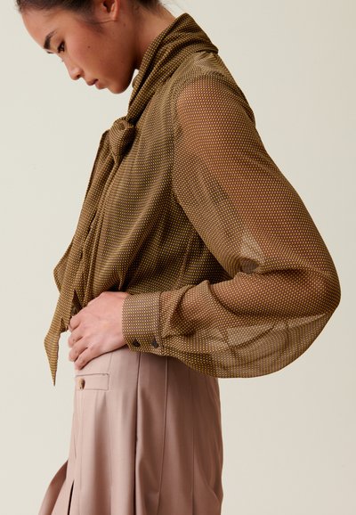 Blusa transparente marrón con un estampado de lunares, mangas largas y cuello atado. Combinada con unos pantalones ajustados de un ligero color beige.