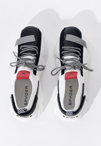 Un paio di sneaker Spyder bianche, nere e rosse con lacci a righe, punte nere e suole bianche spesse, viste dall'alto su una superficie bianca.