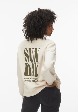 Woman with curly hair wearing a cream sweatshirt with "SUNDAY" and other weekdays printed on the back, paired with black jeans.
