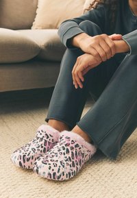 Lepardprint hjemmesko i pink og sort, med blød pink faux pels indvendig og en rund form, hviler på et tekstureret beige tæppe.