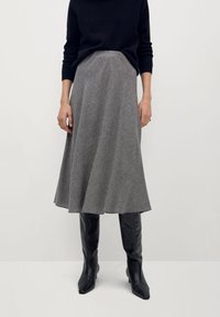 Persona vistiendo una blusa negra de manga larga, una falda midi gris con estampado, y botas de cuero negras hasta la rodilla, de pie contra un fondo sencillo.