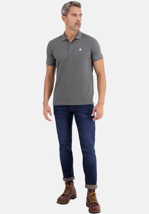 Homme portant un polo gris, un jean bleu foncé retroussé et des bottes marron à lacets, debout et regardant sur le côté.