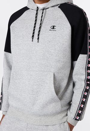 Jasnoszara bluza z kapturem Champion z czarnymi rękawami raglanowymi, logo na piersi i powtarzający się wzór logo na paskach na rękawach, noszona z pasującymi spodniami.