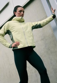 Chaqueta amarilla con cierre en la parte delantera, mangas grises texturizadas y cuello alto. Leggings negros con costuras laterales. Material suave y ligero.