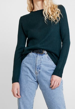 Vrouw met lang blond krullend haar, die een donkergroene gebreide trui draagt, ingestopt in een lichtblauwe high-waisted spijkerbroek met een zwarte riem.