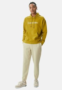 Sudadera amarilla con bolsillo frontal y logo, combinada con pantalones jogger crema que presentan un logo sutil, y zapatillas de color claro con acentos.