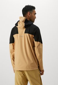 Veste résistante à l'eau dans un design colorblock beige et noir avec une capuche, présentant un logo dans le dos et des manches longues.