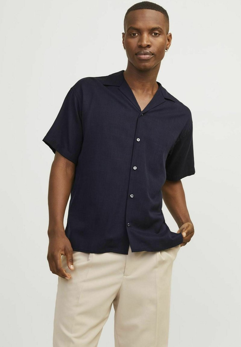 Navy short-sleeve shirt with a collar, button front, and textured fabric. Paired with beige trousers.
