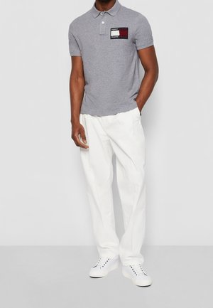 Man draagt een grijs poloshirt met het Tommy Hilfiger Shawn Mendes-logo, witte broek en witte sneakers, staand tegen een witte achtergrond.