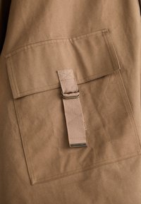 Tissu marron avec une grande poche à rabat rectangulaire et une sangle boucle fixée par un clip en métal. Surface douce et texturée.