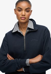 Marineblauw sweatshirt met een ritskraag, met daaronder een gestreepte laag. "Superdry" logo geborduurd op de borst. Lange mouwen met manchetten.