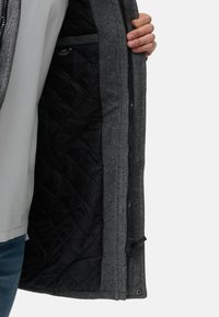 Giacca esterna grigia con trama, fodera trapuntata nera, con chiusura a cerniera e bottoni a pressione. Tasca interna visibile all'interno.