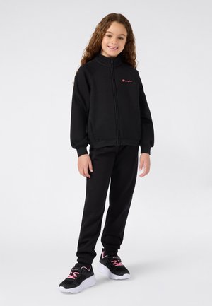 Niña pequeña de pie, vestida con una sudadera negra con cremallera, pantalones negros a juego y zapatillas negras con detalles en rosa, sobre un fondo claro y simple.