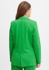 Blazer verde a medida con un ajuste estructurado, solapas de muesca, ventilación trasera y textura suave, usado sobre un atuendo a juego.