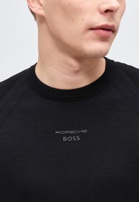 Νεαρό άτομο που φοράει μαύρο πουλόβερ με στρογγυλή λαιμόκοψη, με το λογότυπο "Porsche BOSS" στο κέντρο του στήθους, κοιτάζοντας ελαφρώς προς τα δεξιά.