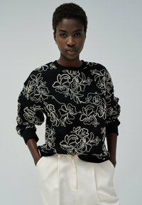 Sweatshirt noir avec broderie florale blanche, col rond et poignets côtelés. Associé à un pantalon taille haute de couleur claire.