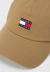Brown baseball cap made of textured fabric, featuring an embroidered logo with navy, white, and red accents. Curved brim and stitched detailing.
