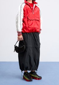 Person som bär en röd och vit jacka, svart cargokjol med dragsko i nederkant, svarta och röda sneakers, och håller en liten svart handväska.