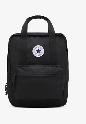 Sac à dos carré noir avec poche zippée à l'avant, deux poignées et un patch rond du logo Converse All Star centré près du haut.