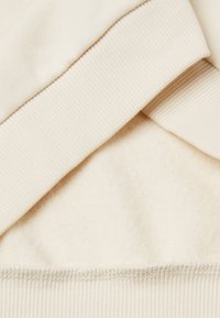 Beżowy materiał sweatshirtu z miękkim, polarowym wnętrzem. Posiada ściągacze na mankietach i gładką powierzchnię. Szczegóły szwów widoczne wzdłuż dolnej krawędzi.
