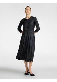 Schwarzes, plissiertes Kleid mit langen Ärmeln und Rundhalsausschnitt, aus einem glatten, glänzenden Material gefertigt, kombiniert mit schwarzen Schuhen mit Schleifen.