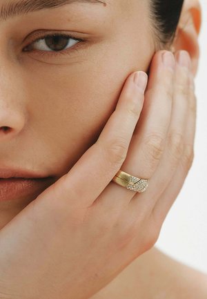 Primer plano del rostro de una mujer con la mano cubriendo parcialmente la mejilla, llevando un anillo de oro con un conjunto de pequeños diamantes.