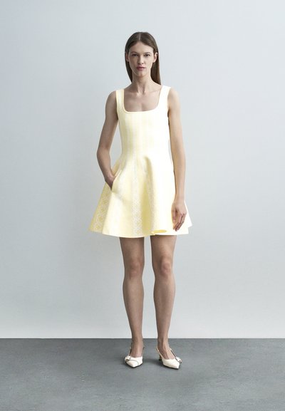 Γυναίκα που φοράει αμάνικο παλαιό κίτρινο φόρεμα με διακριτικά σχέδια και τσέπες, στέκεται μπροστά σε έναν απλό ανοιχτό γκρι φόντο.